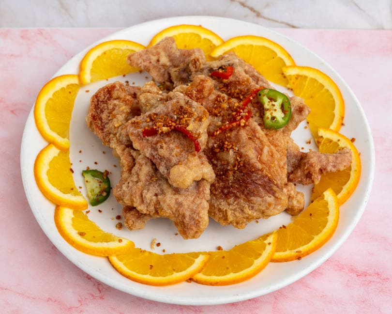 Crispy fried chicken pieces garnished with red and green chilies, surrounded by orange slices on a white plate.