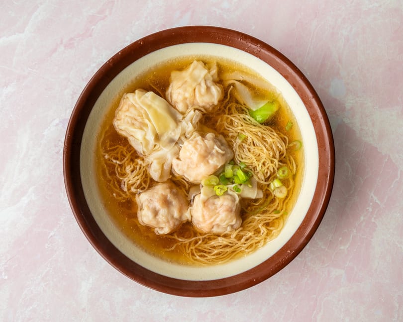 Wonton noodle soup with five wontons, thin noodles, green onions, and bok choy in a brown bowl.