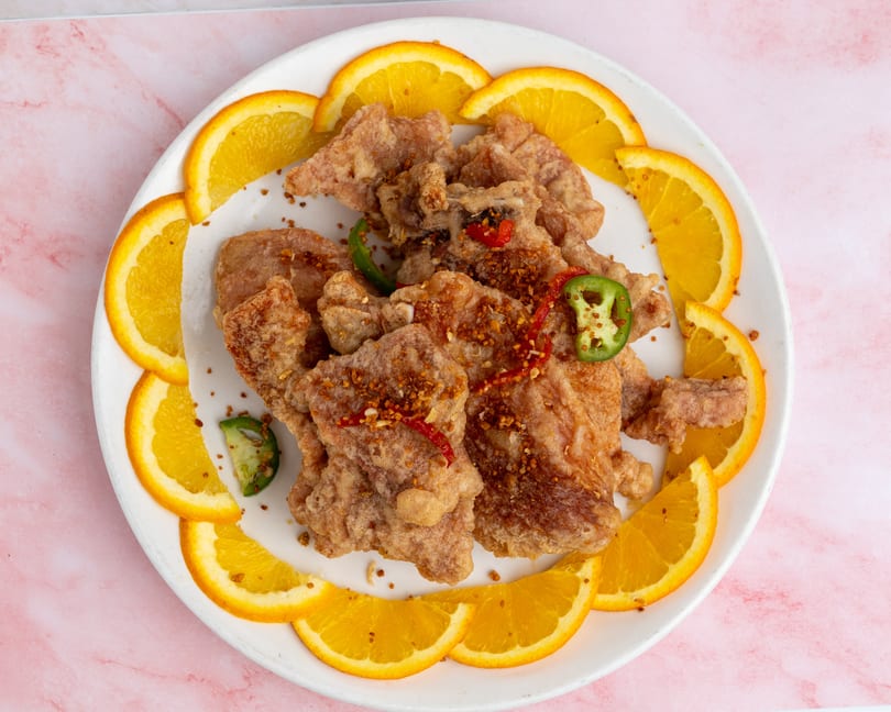 Crispy fried chicken pieces garnished with red and green peppers, surrounded by orange slices on a white plate.