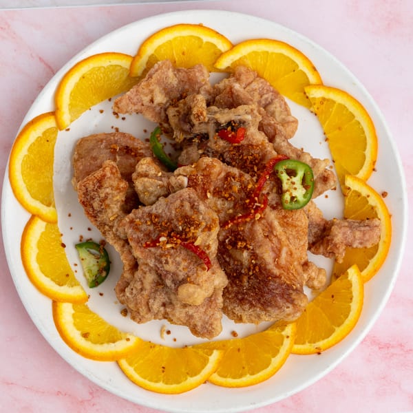 Crispy fried chicken pieces garnished with red and green peppers, surrounded by orange slices on a white plate.