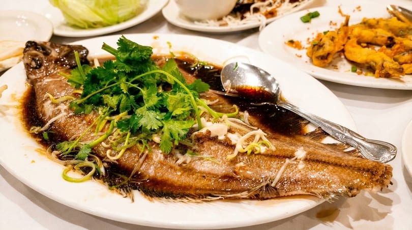 Steamed whole fish with dark sauce, cilantro, and scallions on a white plate with a spoon.