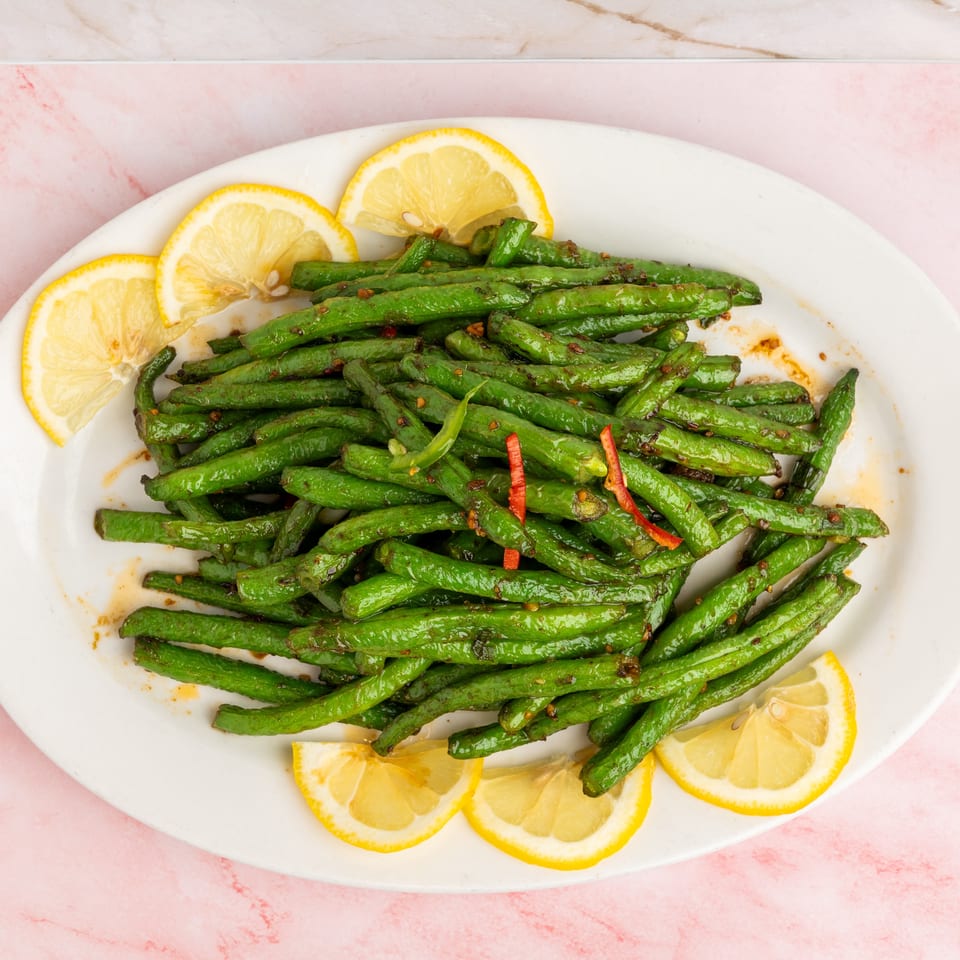 Sautéed green beans with red chili flakes and lemon slices on a white oval plate.