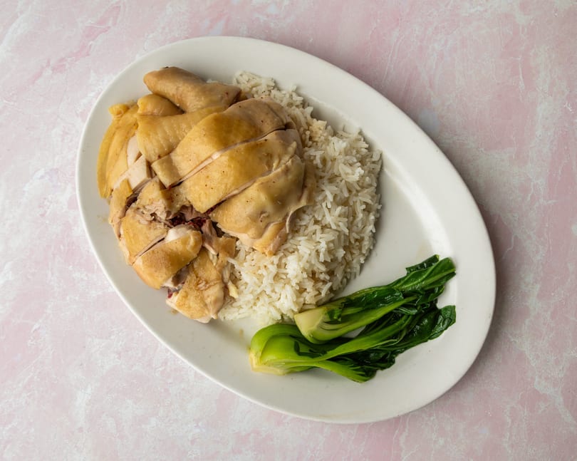 Hainanese chicken rice with sliced poached chicken, white rice, and green bok choy on a white plate.