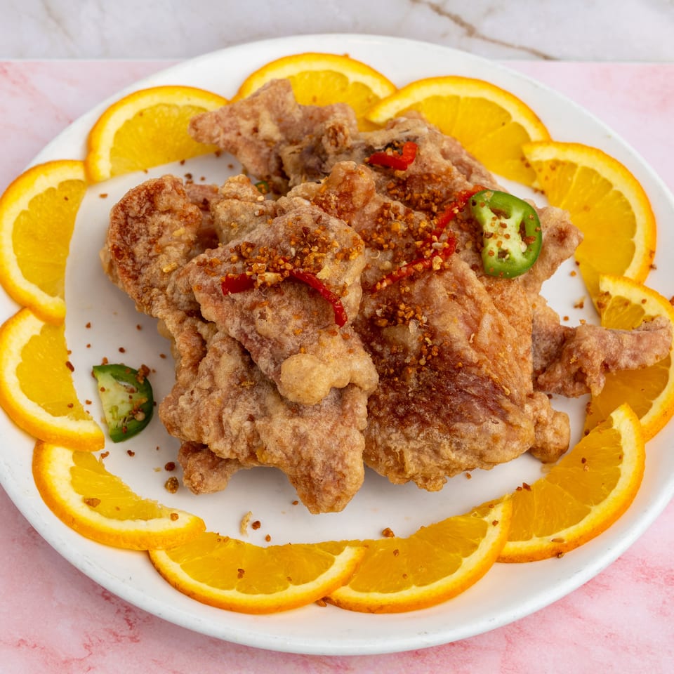 Crispy fried chicken pieces garnished with red and green chilies, surrounded by orange slices on a white plate.