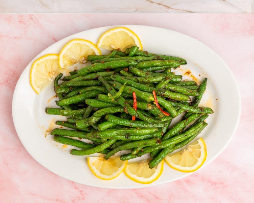 Sautéed green beans with red chili flakes and lemon slices on a white oval plate.