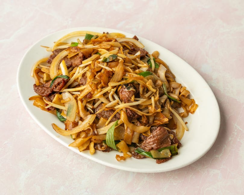A white oval plate filled with beef chow fun, featuring wide noodles, beef, onions, and bean sprouts.